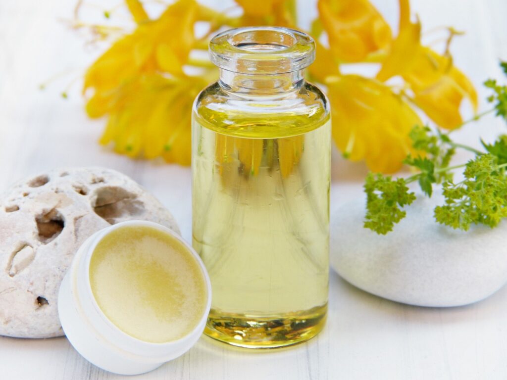 Small jar of beeswax lip balm and apothecary bottle of herbal infused body oil surrounded by fresh flower and herbs.